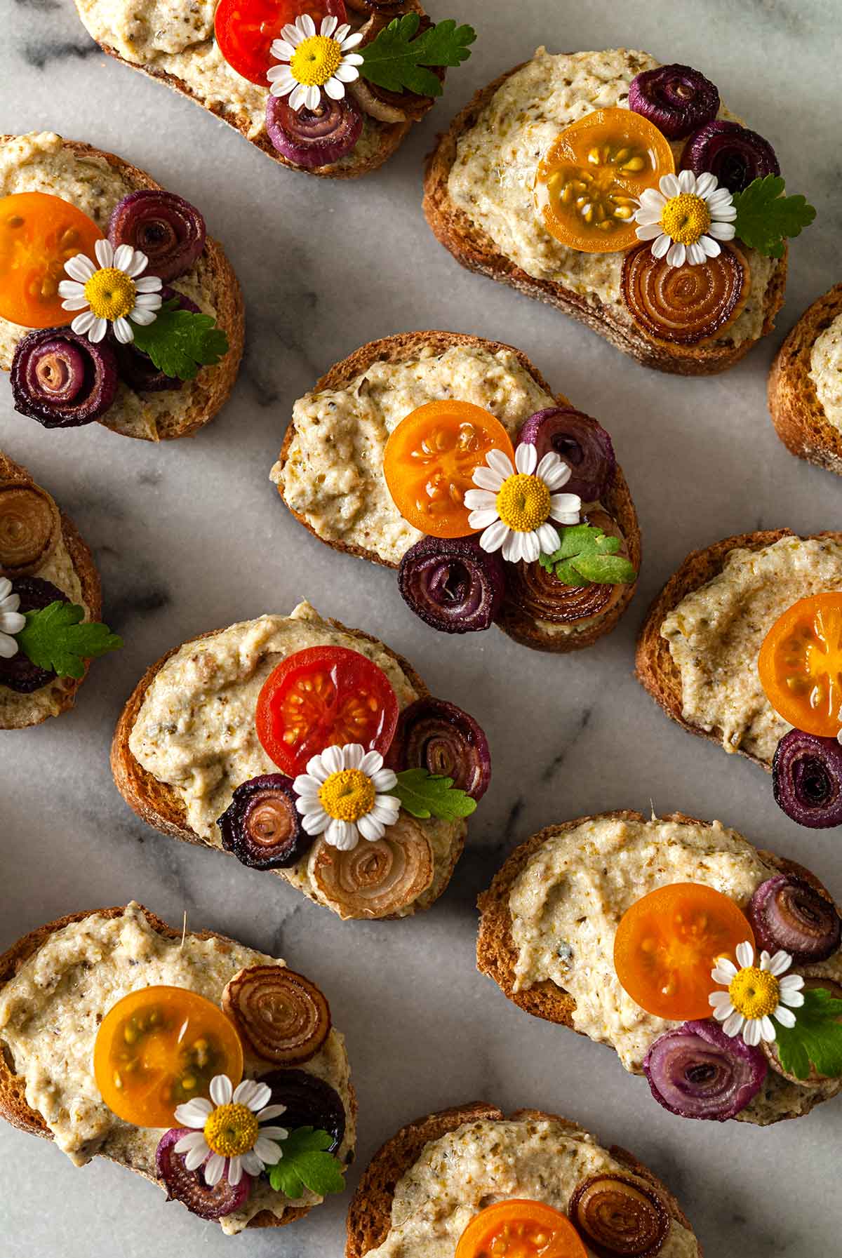 11 crostini garnished with daisies, onions and tomatoes on a marble plate.