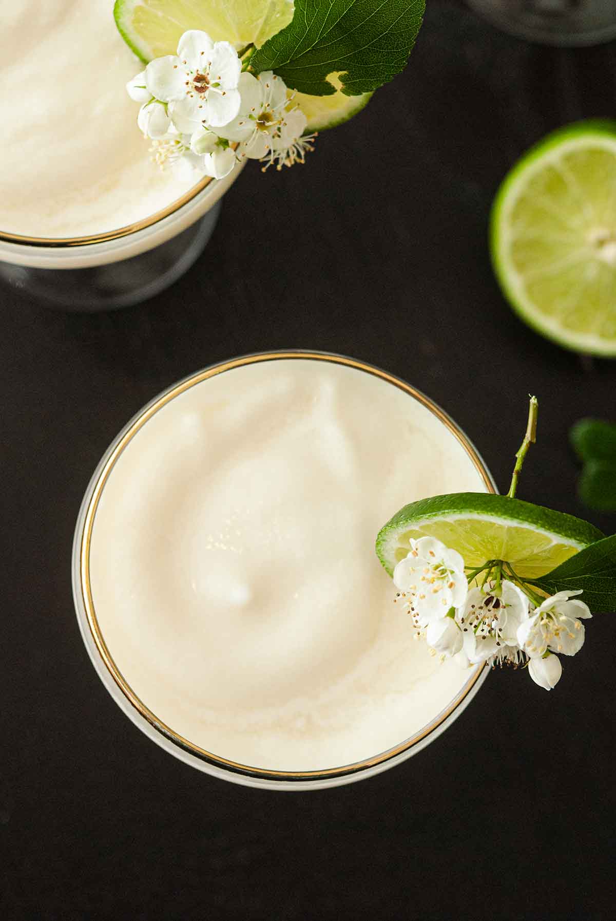 The tops of 2 piña coladas with lime-flower garnishes on a table with a slice of lime.