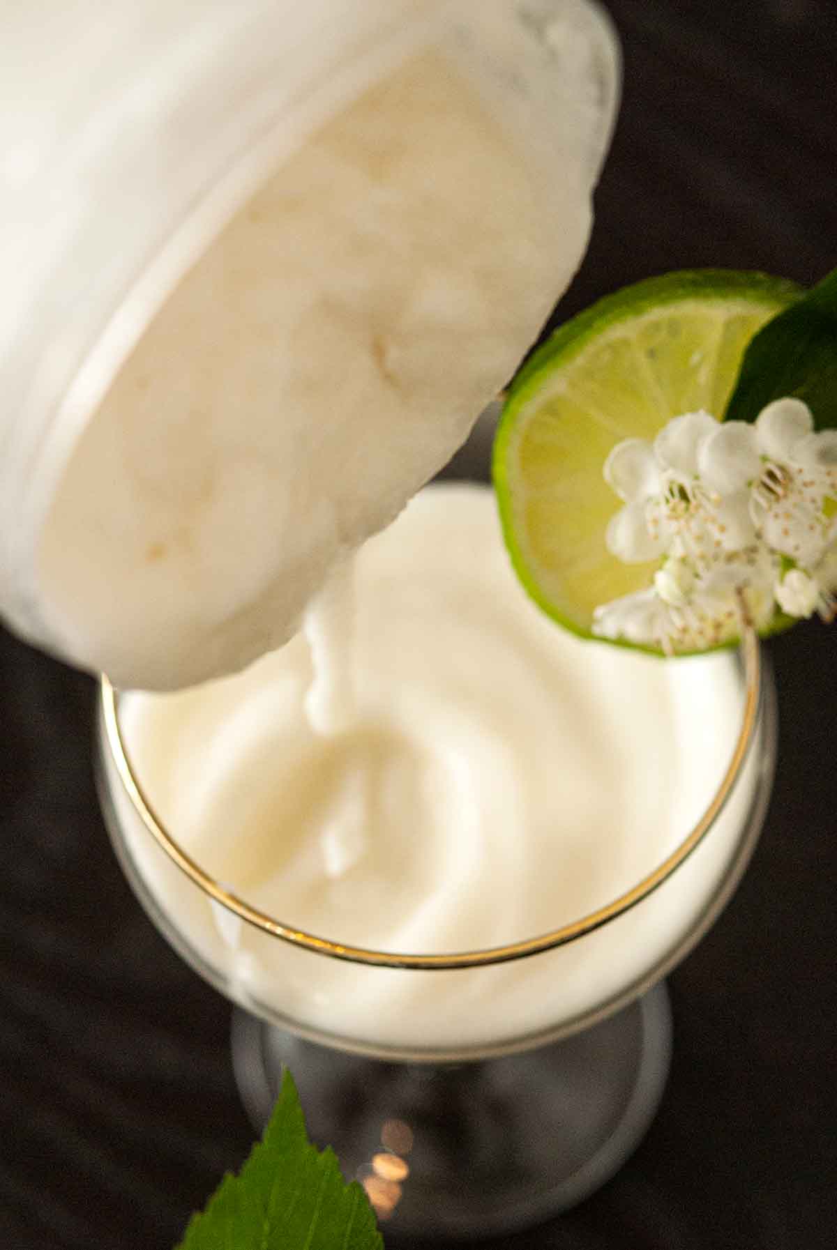 A piña colada cocktail being poured out of a container into a garnished coup glass on a table.