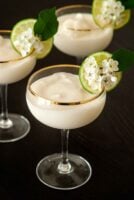 3 piña coladas in coup glasses, garnished with a lime and flowers on a table.