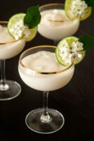 3 piña coladas in coup glasses, garnished with a lime and flowers on a table.