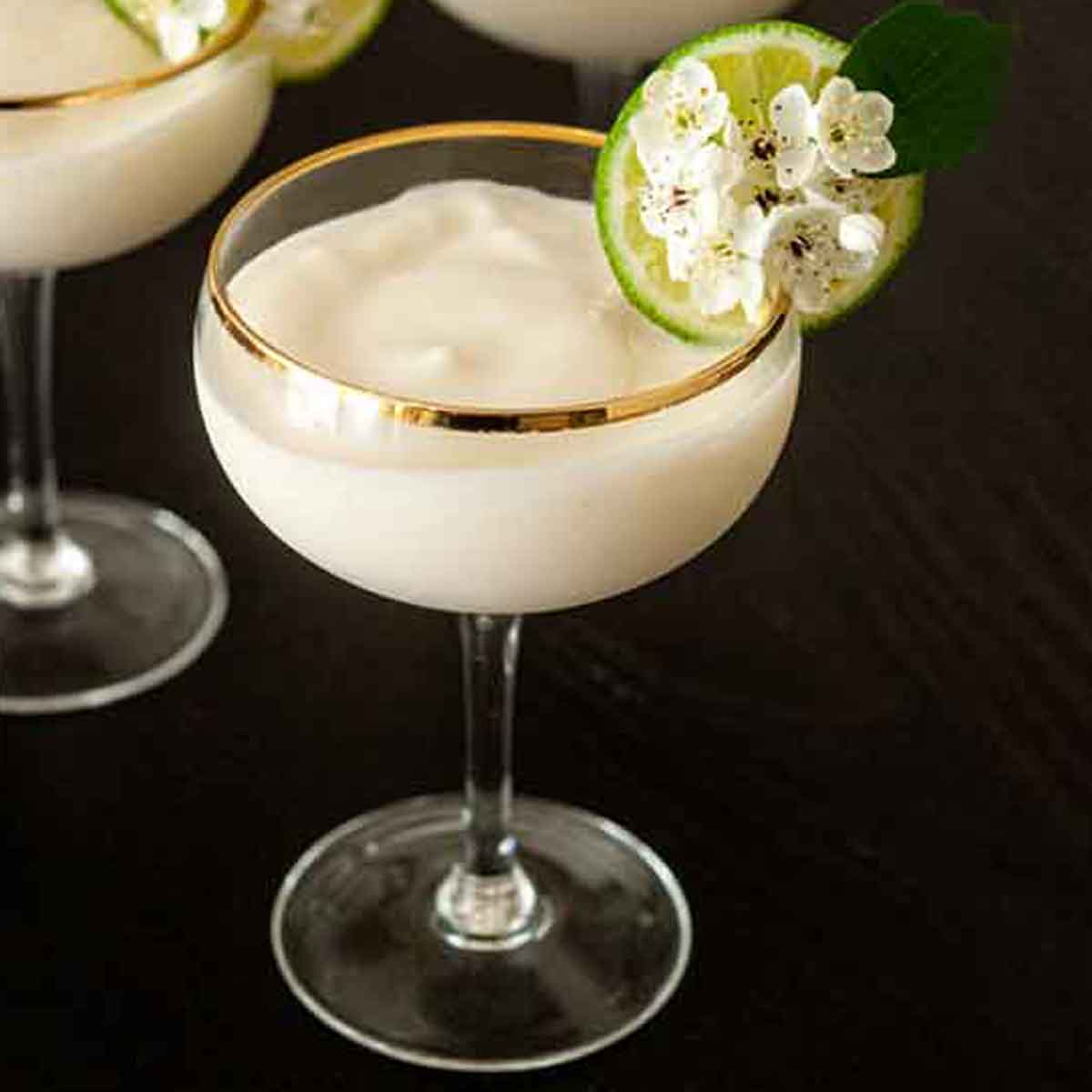 A piña colada in coup glasses, garnished with a lime and flowers on a table.
