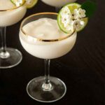 A piña colada in a coup glass, garnished with a lime and flowers.