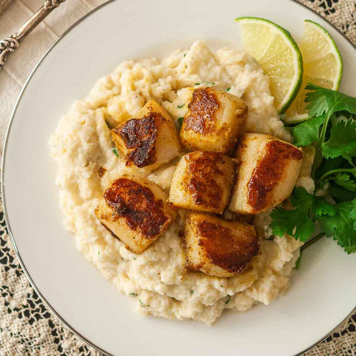 6 curry-seared scallops on a plate with mashed cauliflower, parsley and 2 lime slices.
