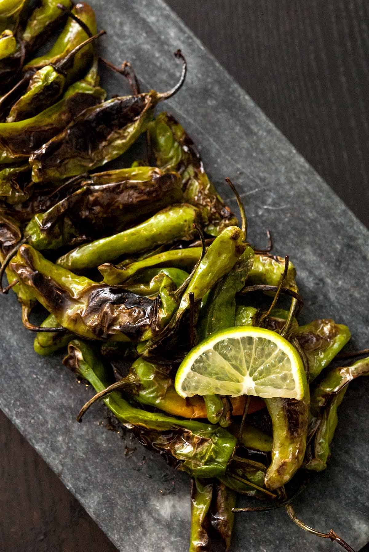 Blistered shishito peppers on a slate with a slice on lime.