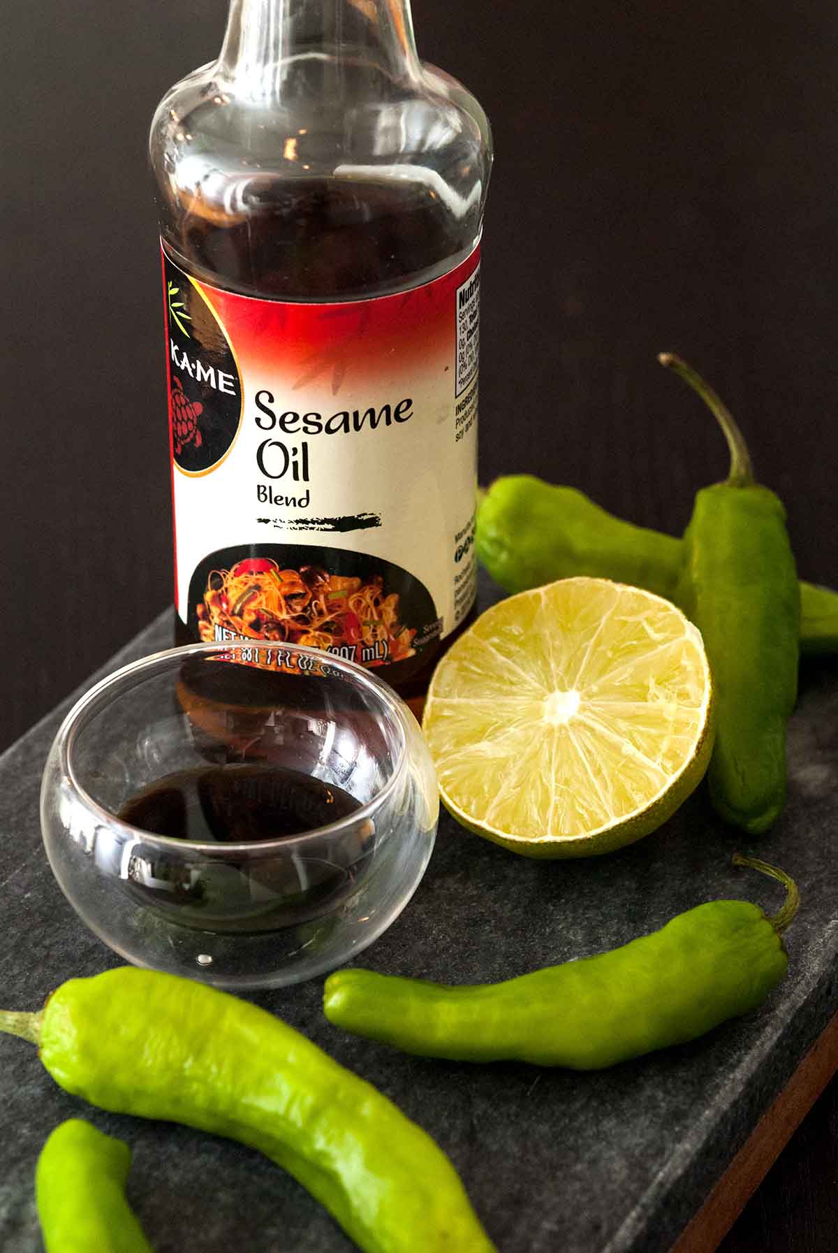 A bottle of sesame oil, a bowl of soy sauce, a lime slice and shishito peppers scattered on a slate.