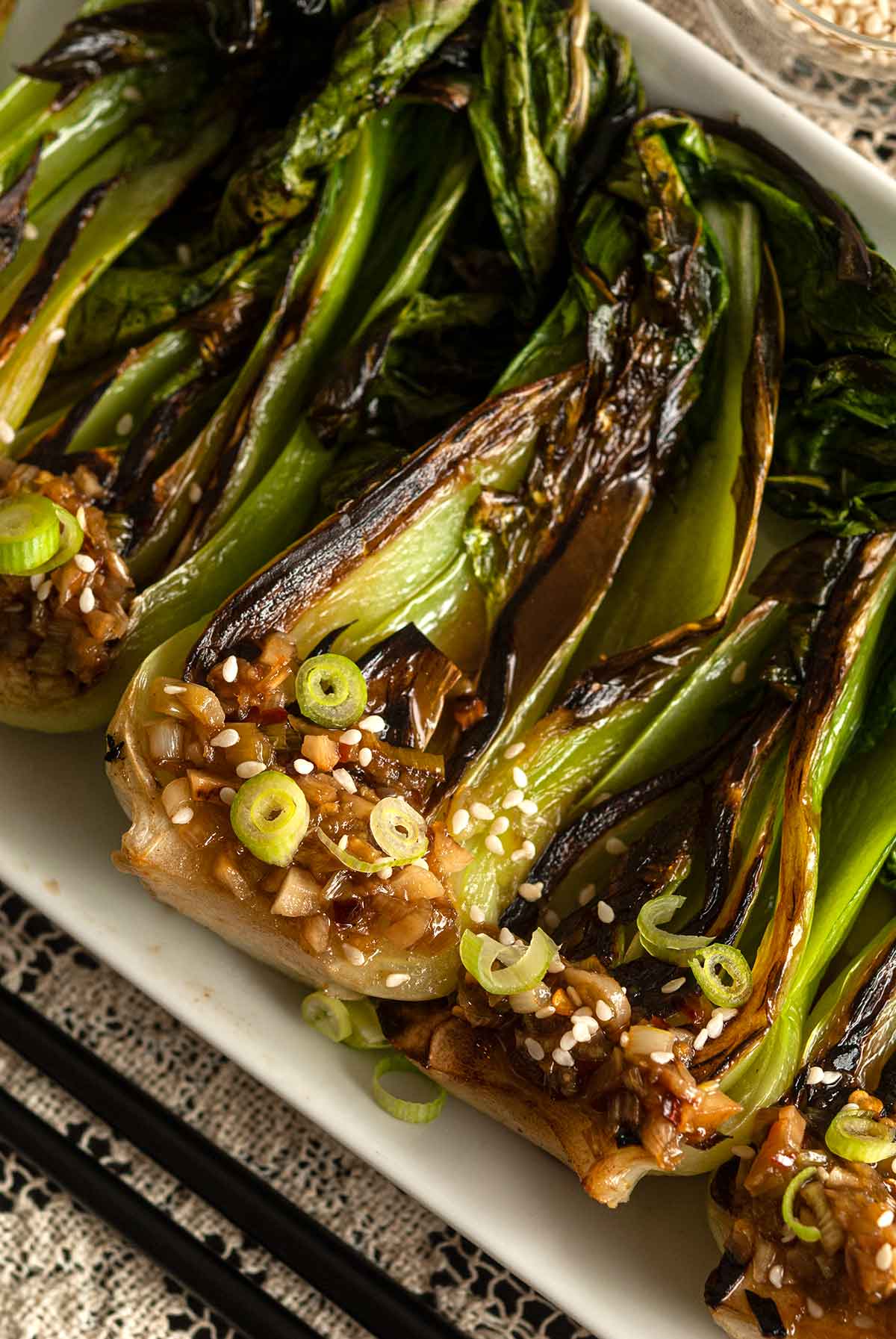 Seared baby bok choy on a plate beside chop sticks on a lace table cloth.