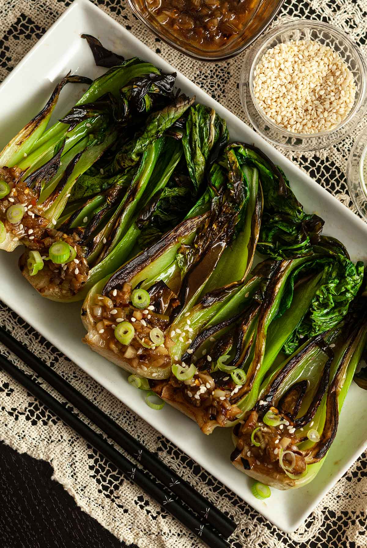 5 seared baby bok choy on a plate beside chop sticks, a bowl of sesame seeds and sauce on a lace table cloth.