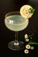 A Lemon Drop cocktail, garnished with a lemon slice and 3 small daisies on a table, with a bouquet in the background.