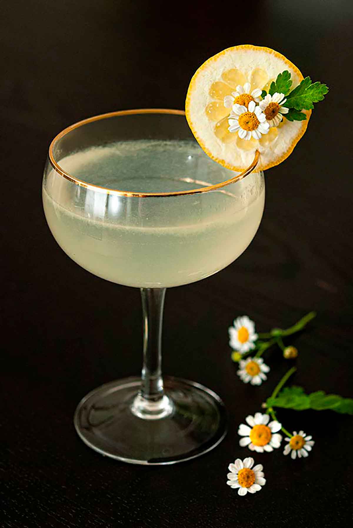 A Lemon Drop cocktail, garnished with a lemon slice and 3 small daisies on a table, with a bouquet in the background.