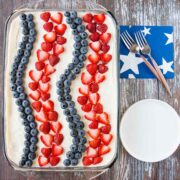 A cake decorated with strawberries and blueberries in stripes on a wooden table, beside 2 forks, napkins and plates.