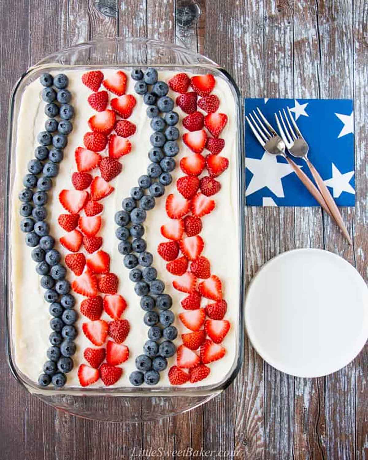 A cake decorated with strawberries and blueberries in stripes on a wooden table, beside 2 forks, napkins and plates.