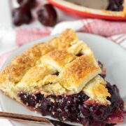 A slice of cherry pie on a plate with a fork, in front of a pie tin.