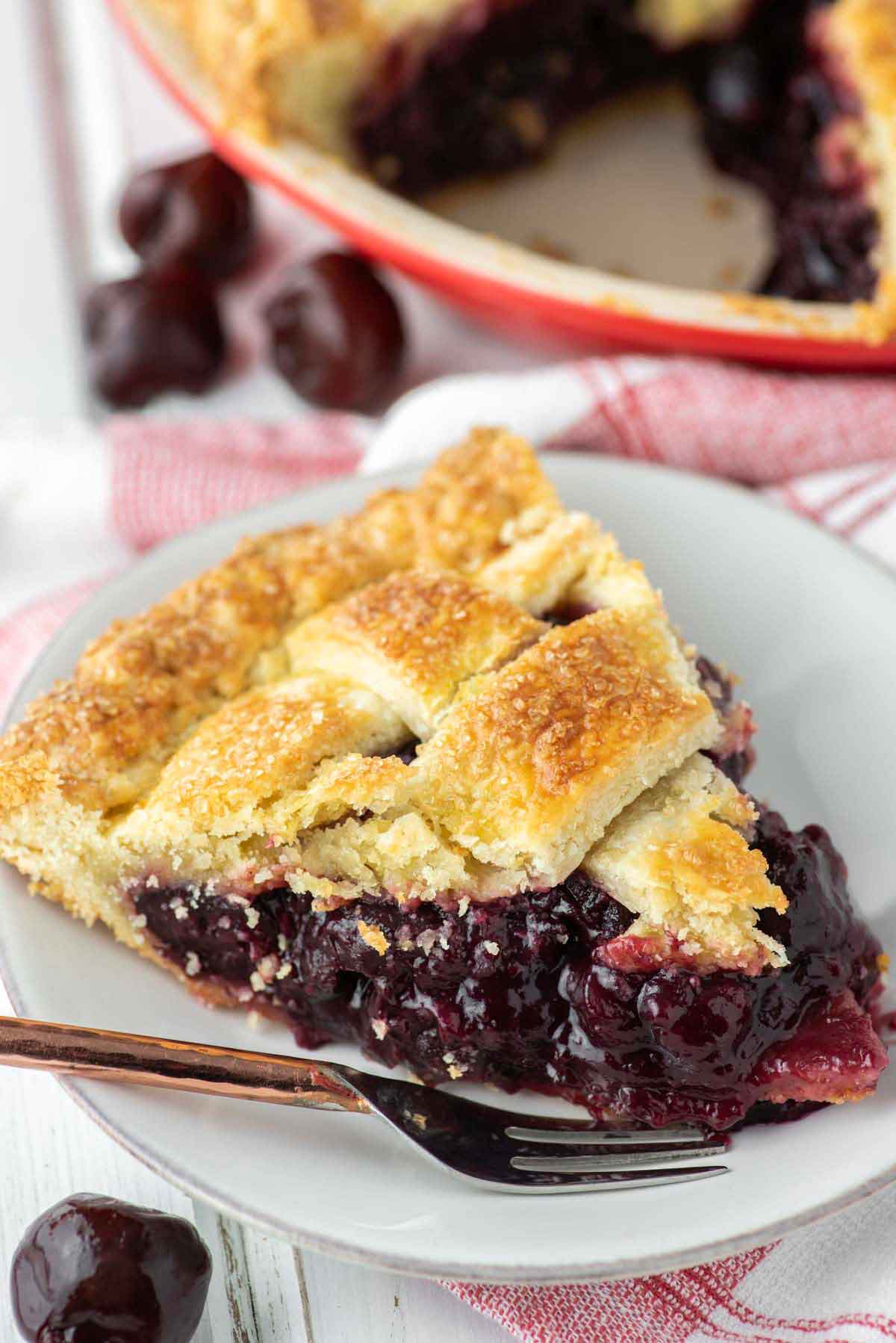 A slice of cherry pie on a plate with a fork, in front of a pie tin.