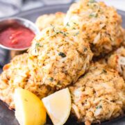 About 6 crab cakes on a plate with sliced lemon and a bowl of cocktail sauce.