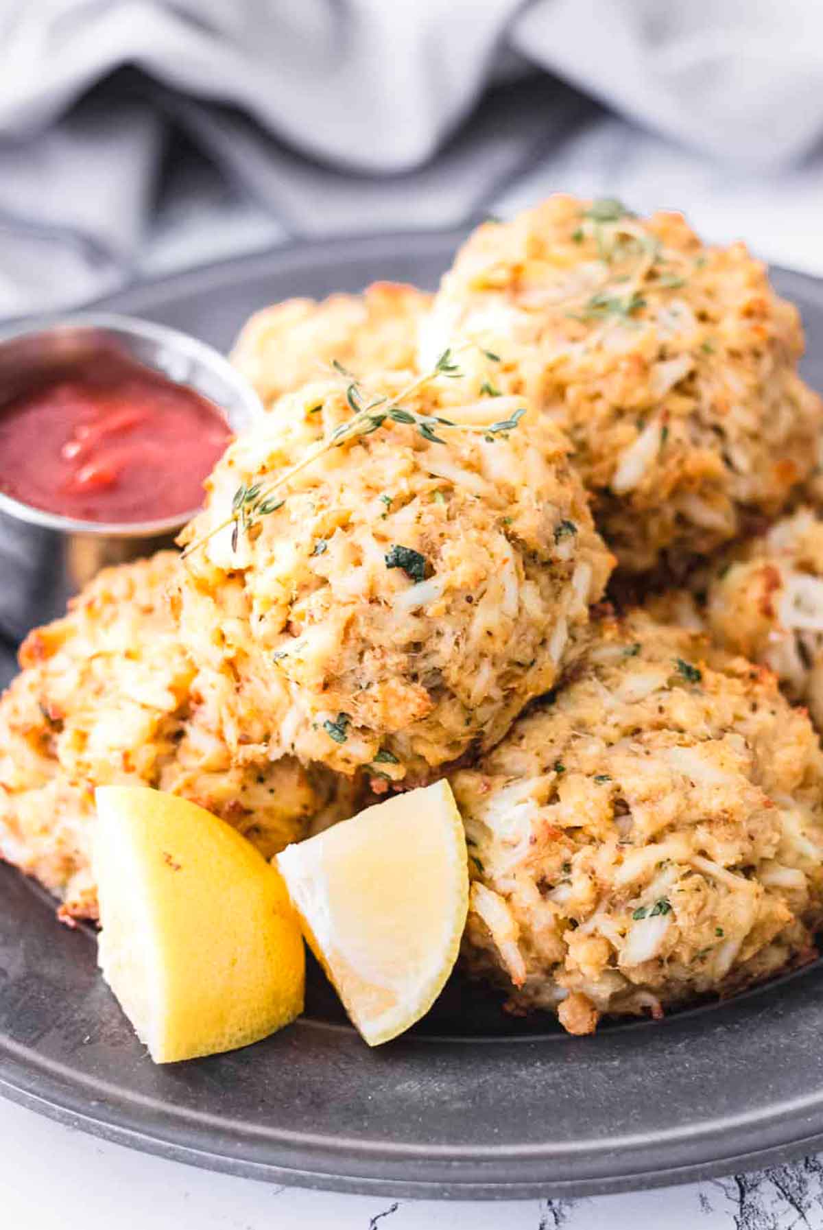 About 6 crab cakes on a plate with sliced lemon and a bowl of cocktail sauce.