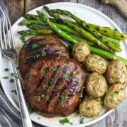 2 grilled mushroom caps on a plate with potatoes and asparagus on the side, on a table.