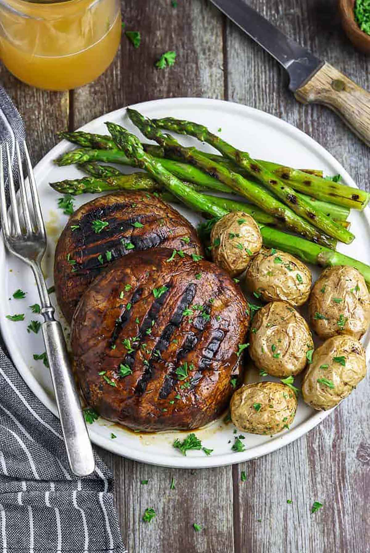 2 grilled mushroom caps on a plate with potatoes and asparagus on the side, on a table.