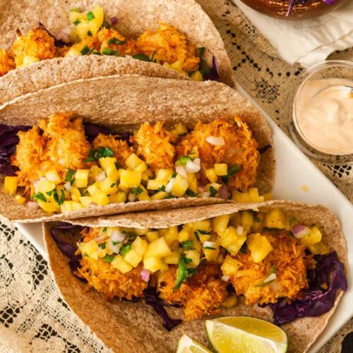 3 coconut shrimp tacos on a plate with pineapple salsa beside 2 lime slices, on a lace tablecloth.