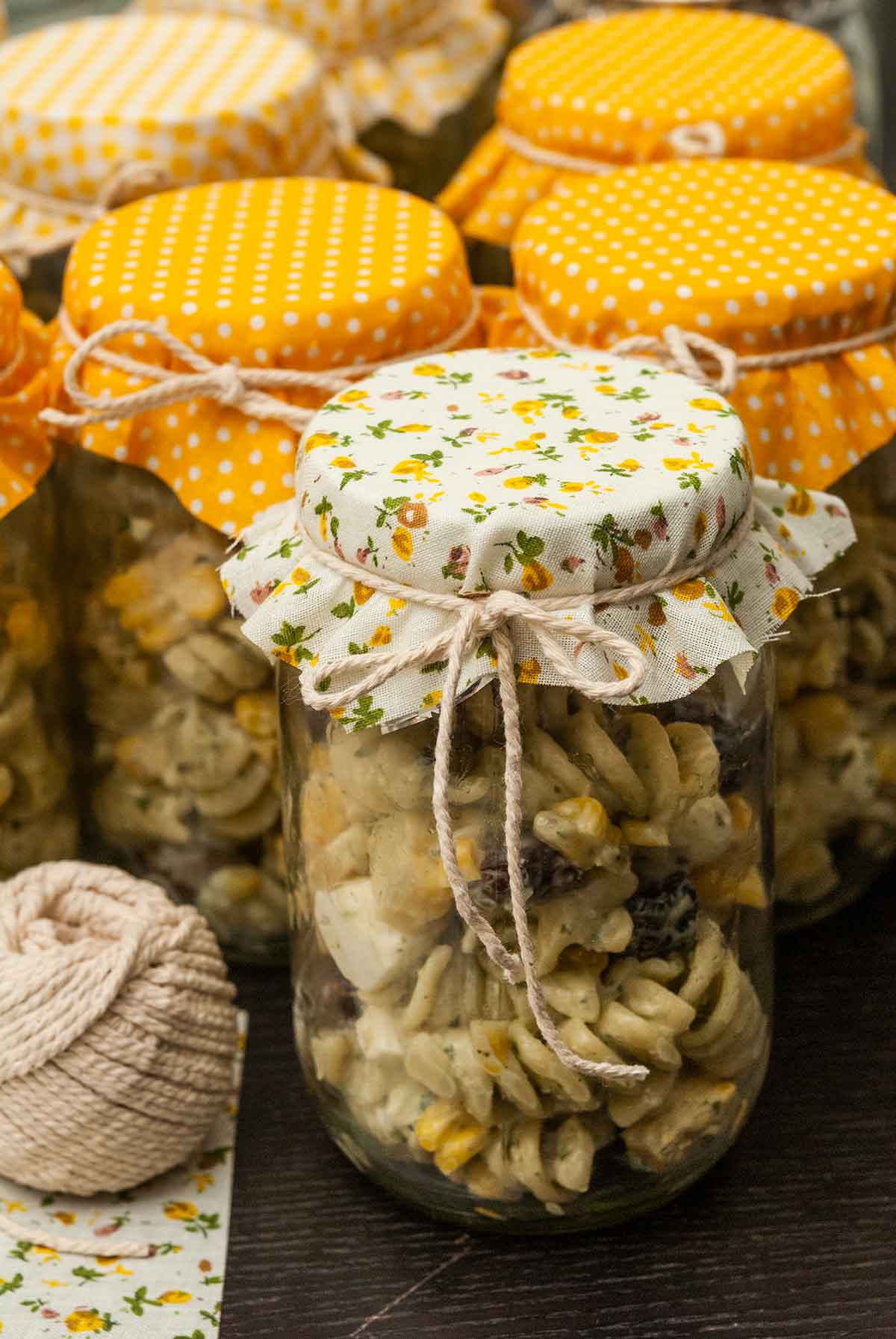 6 jars of pasta salad on a table, topped with fabric and tied with string.