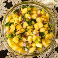 Pineapple jalapeño salsa in a small bowl on a lace tablecloth, beside a spoon and 2 small bunches of flowers.