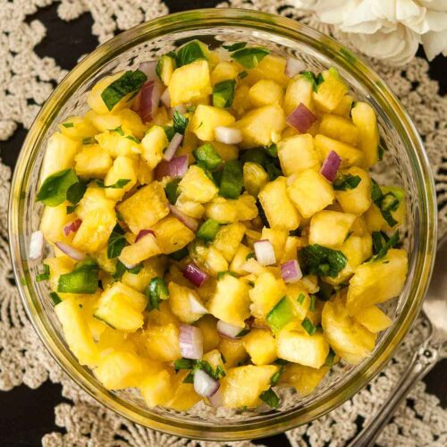 Pineapple jalapeño salsa in a small bowl on a lace tablecloth, beside a spoon and 2 small bunches of flowers.