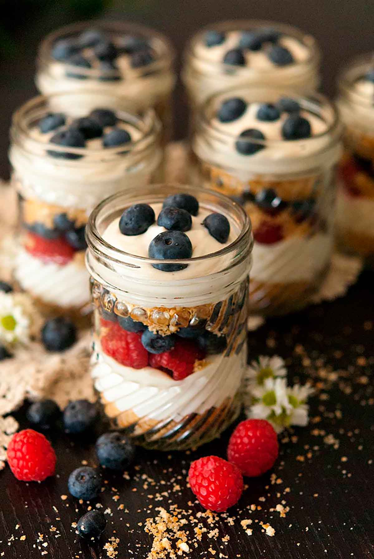 6 mason jars with cheesecake mouse, cookie crumbles and berries on a black table, sprinkled with cookie crumbs and berries.