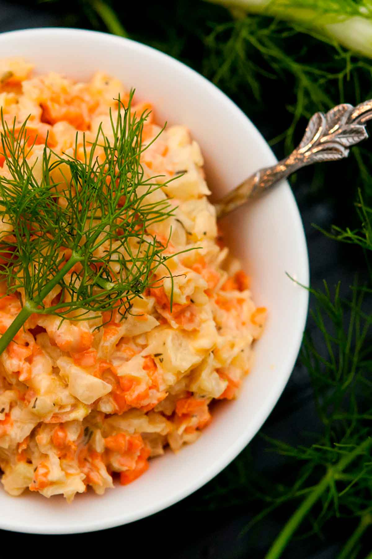 A bowl of fennel & carrot coleslaw in a small white bowl, garnished with a fennel frond next to larger fronds on a table.