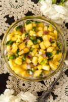 Pineapple jalapeño salsa in a small bowl on a lace tablecloth, beside a spoon and 2 small bunches of flowers.