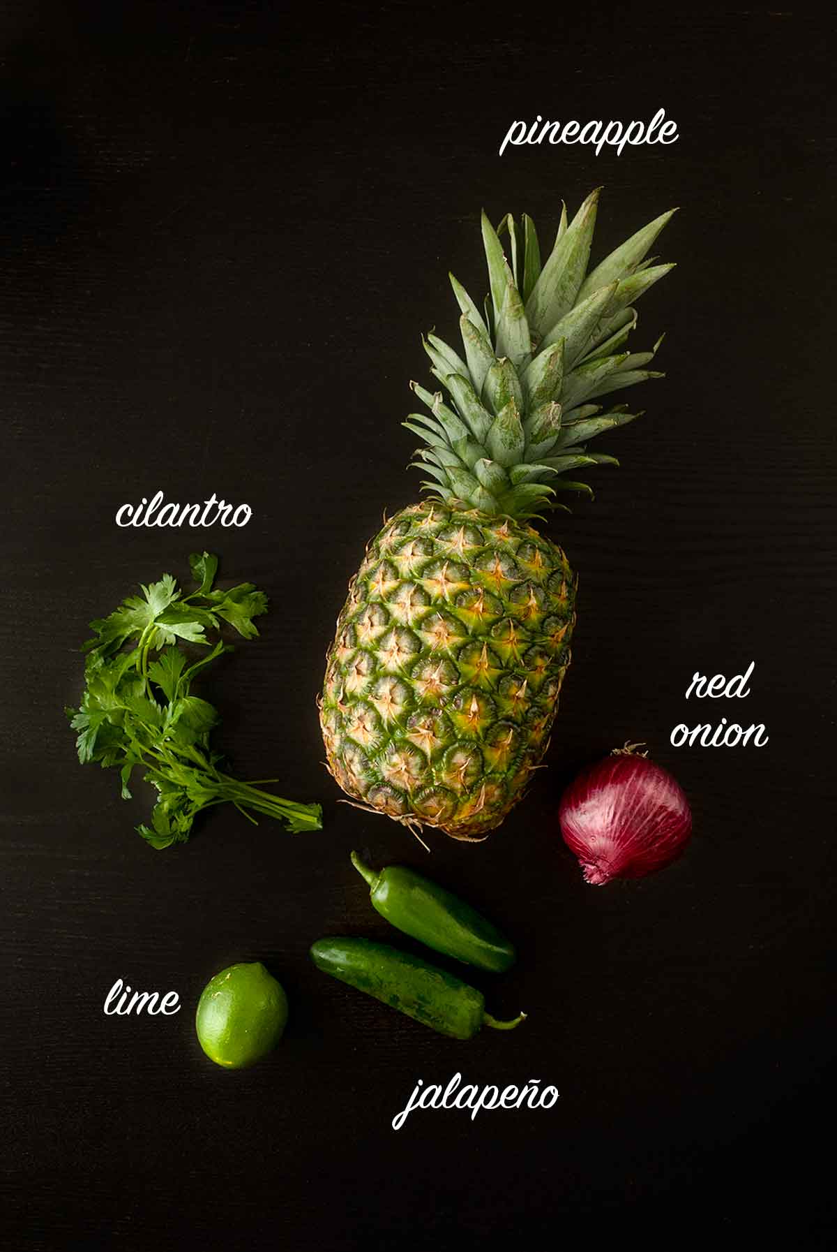 Ingredients for making pineapple jalapeño salsa on a black table with titles describing what they are.