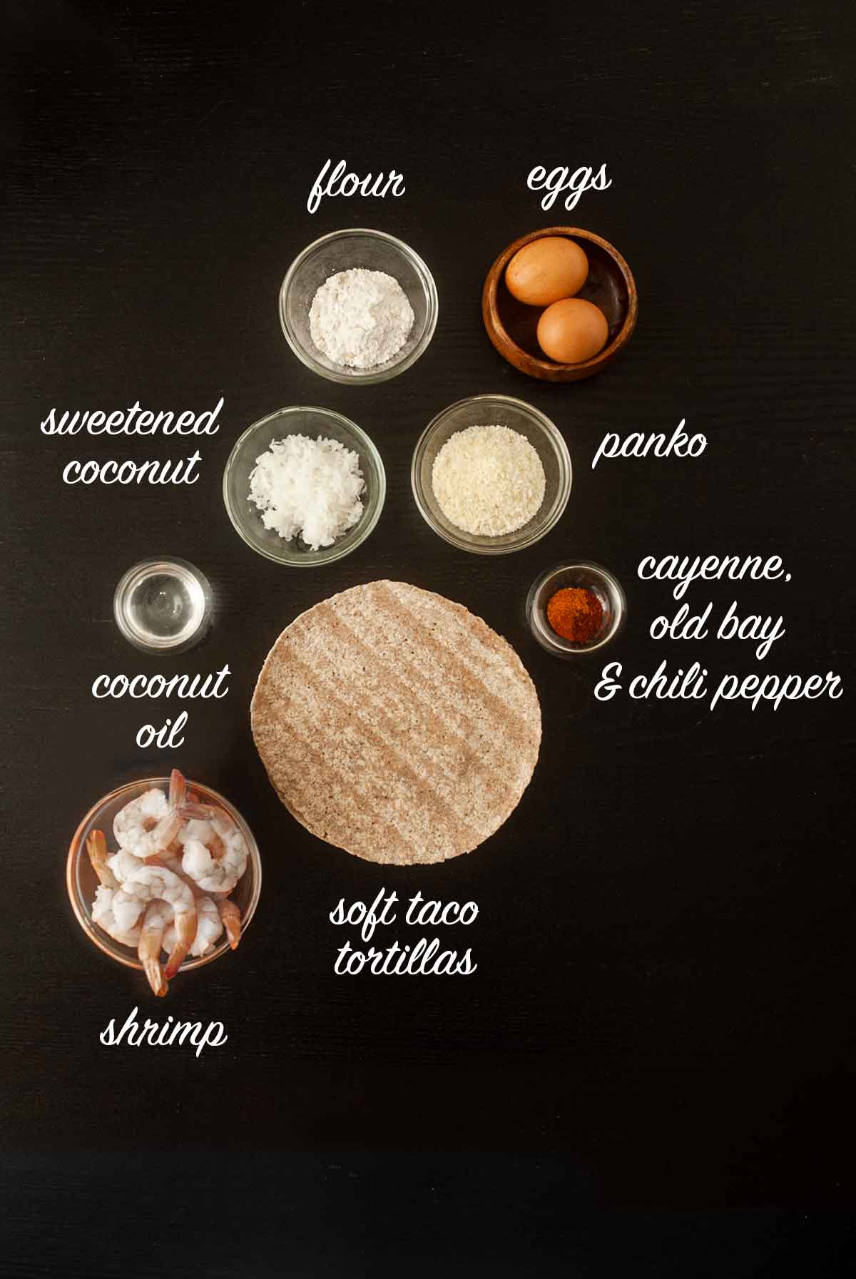 A number of ingredients for making coconut shrimp tacos on a table with titles describing what they are.