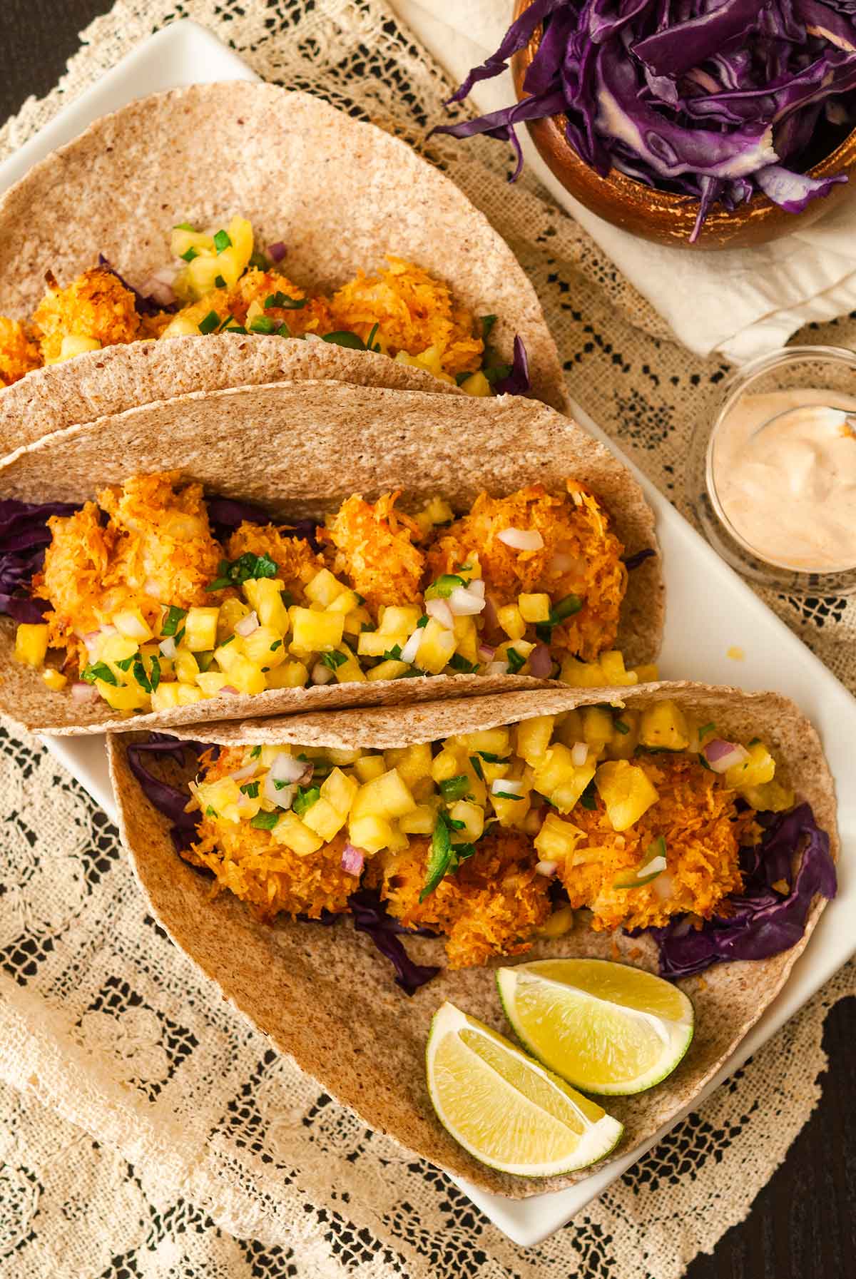 3 coconut shrimp tacos on a plate with pineapple salsa beside 2 lime slices, on a lace tablecloth.