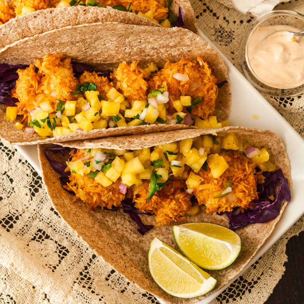 2 coconut shrimp tacos on a lace table cloth table garnished with pineapple salsa with a side of 2 lime slices.