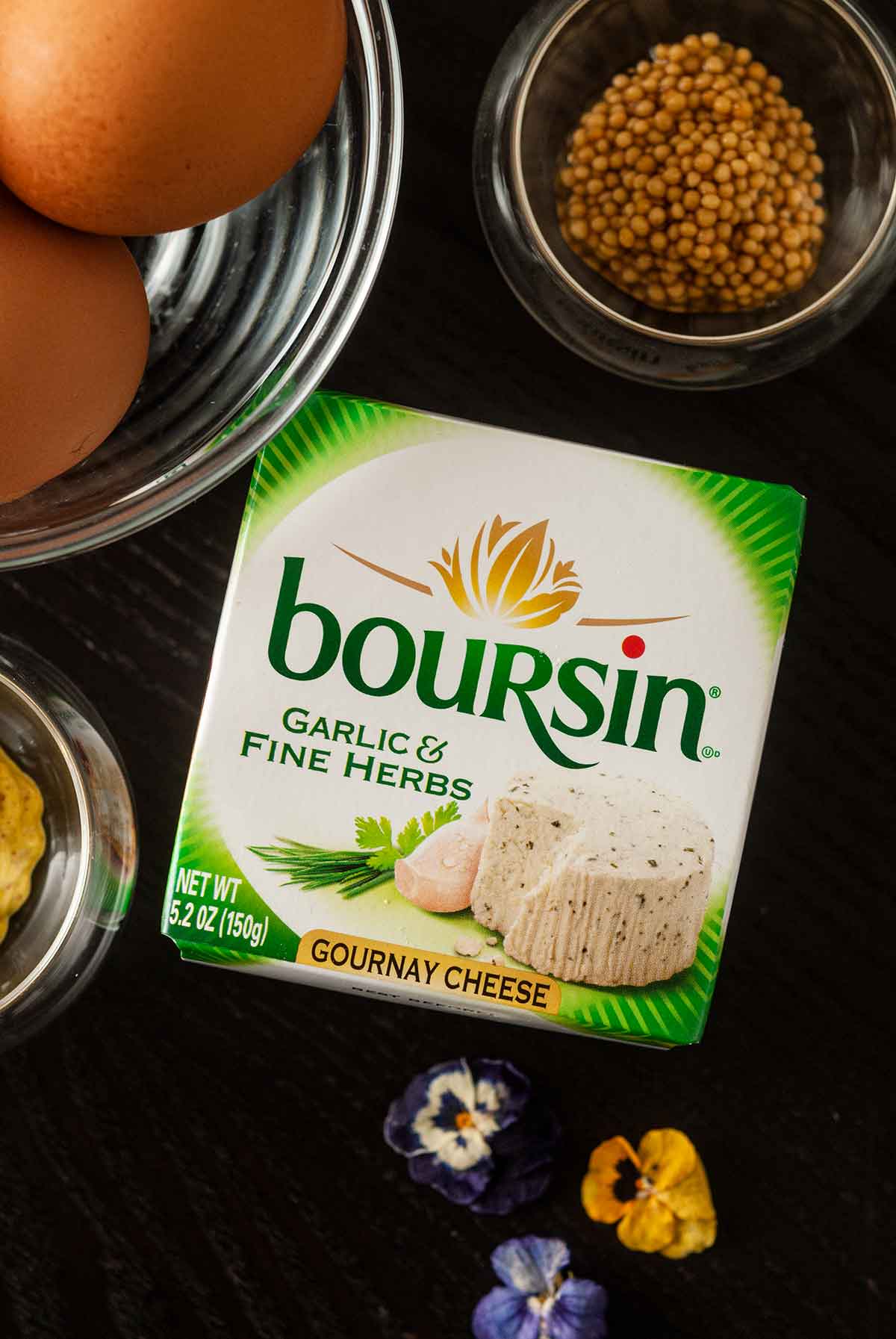 A package of Boursin cheese on a table beside a bowl of eggs, mustard caviar and a few scattered flowers.