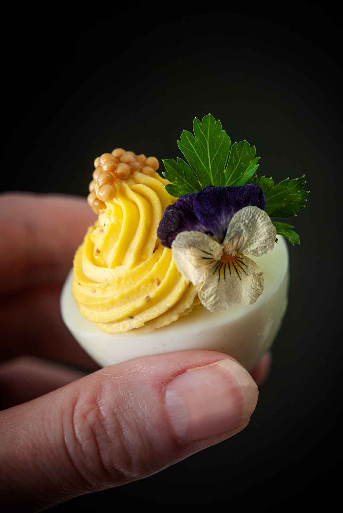 Fingers holding a deviled egg, garnished with a flower, cilantro and mustard caviar.