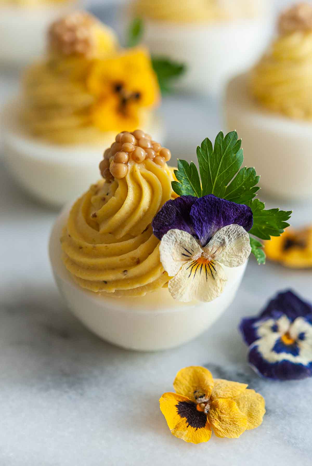 4 deviled eggs on marble, garnished with flowers, cilantro and mustard caviar beside 3 scattered flowers.