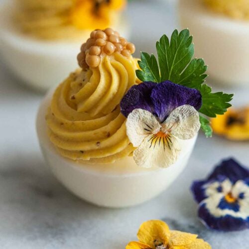 1 deviled egg on marble, garnished with flowers, cilantro and mustard caviar beside 3 scattered flowers.