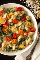 A bowl of gnocchi with tomatoes and spinach on a table beside flowers and a spoon on a napkin.