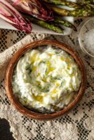 A bowl of tzatziki, drizzled with olive oil, on a lace table cloth beside vegetables.