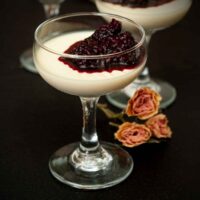 A glass of panna cotta with berry sauce on a table beside 3 dry roses.