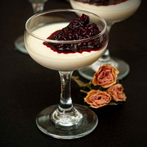 A glass of panna cotta with berry sauce on a table beside 3 dry roses.