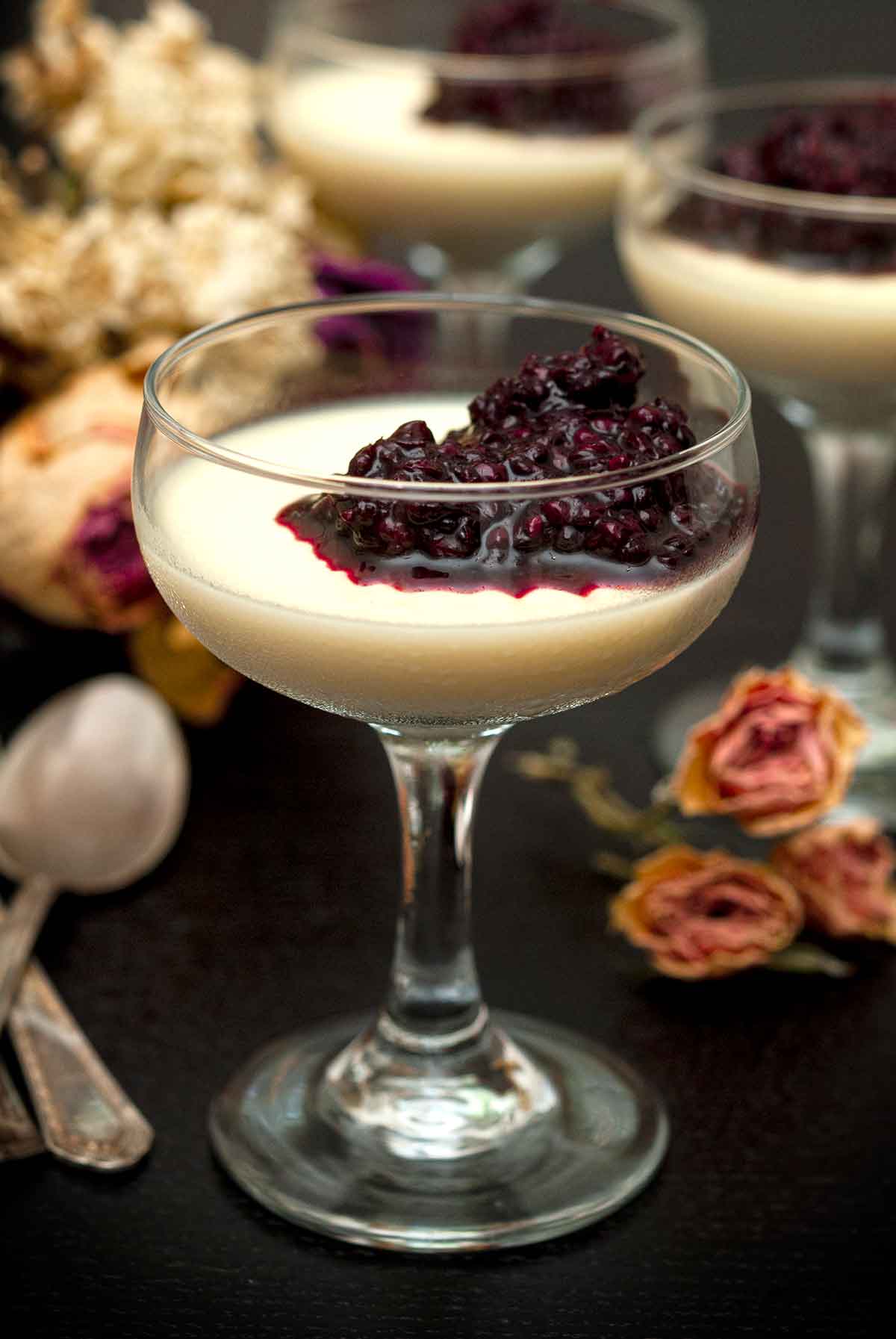 3 glasses of panna cotta with berry sauce on a table with flowers and spoons.