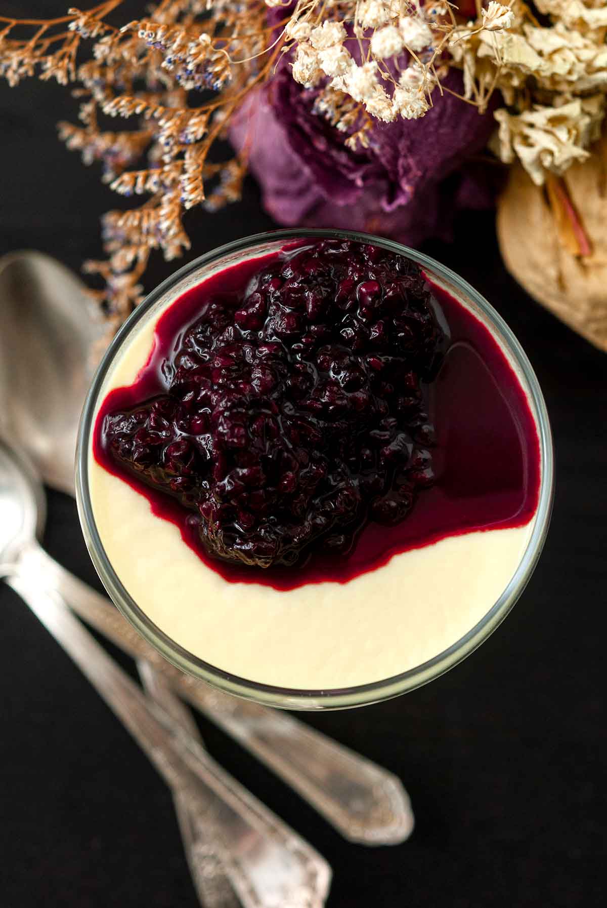 The top of a panna cotta in a glass with berry sauce, surrounded by flowers and 3 spoons below.