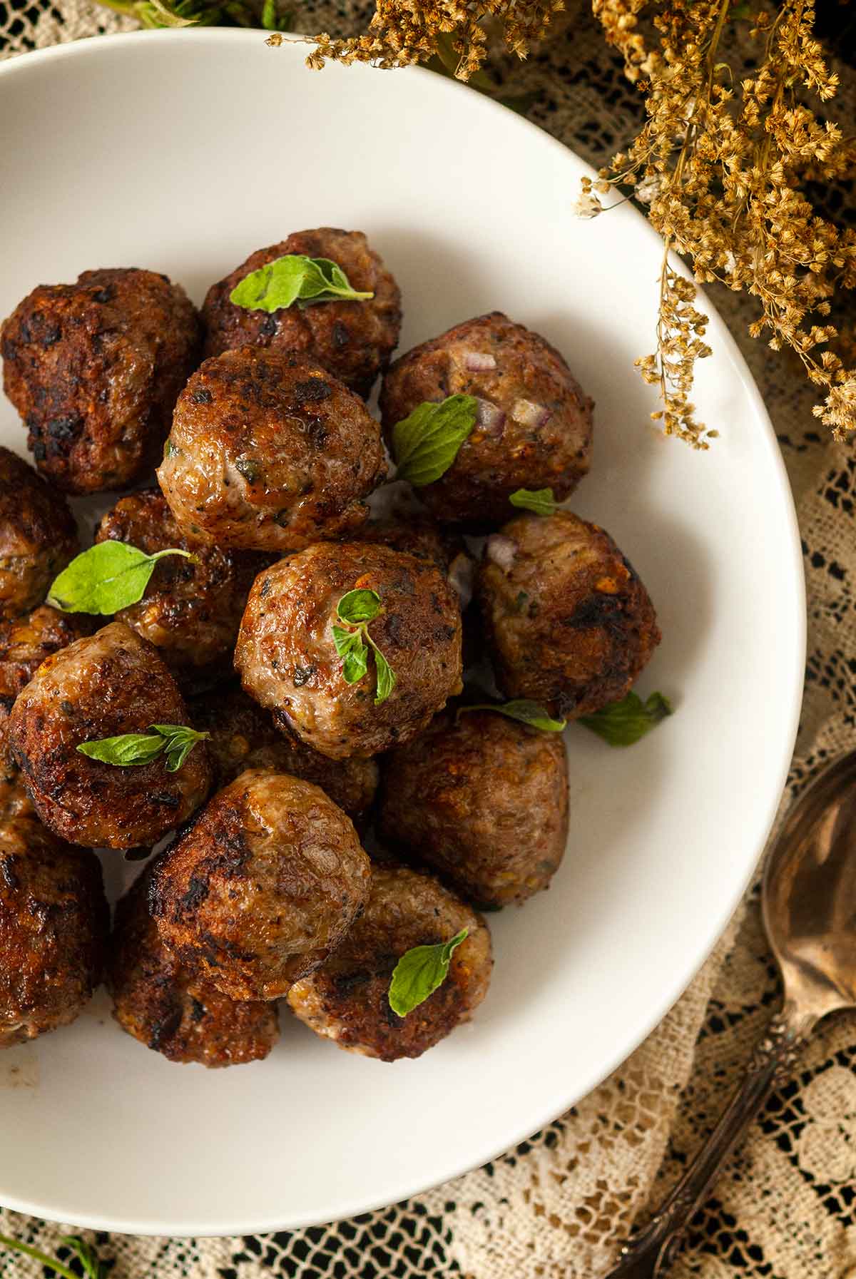 About 15 meatballs in a bowl, sprinkled with fresh oregano, surrounded by wispy flowers on a lace tablecloth.