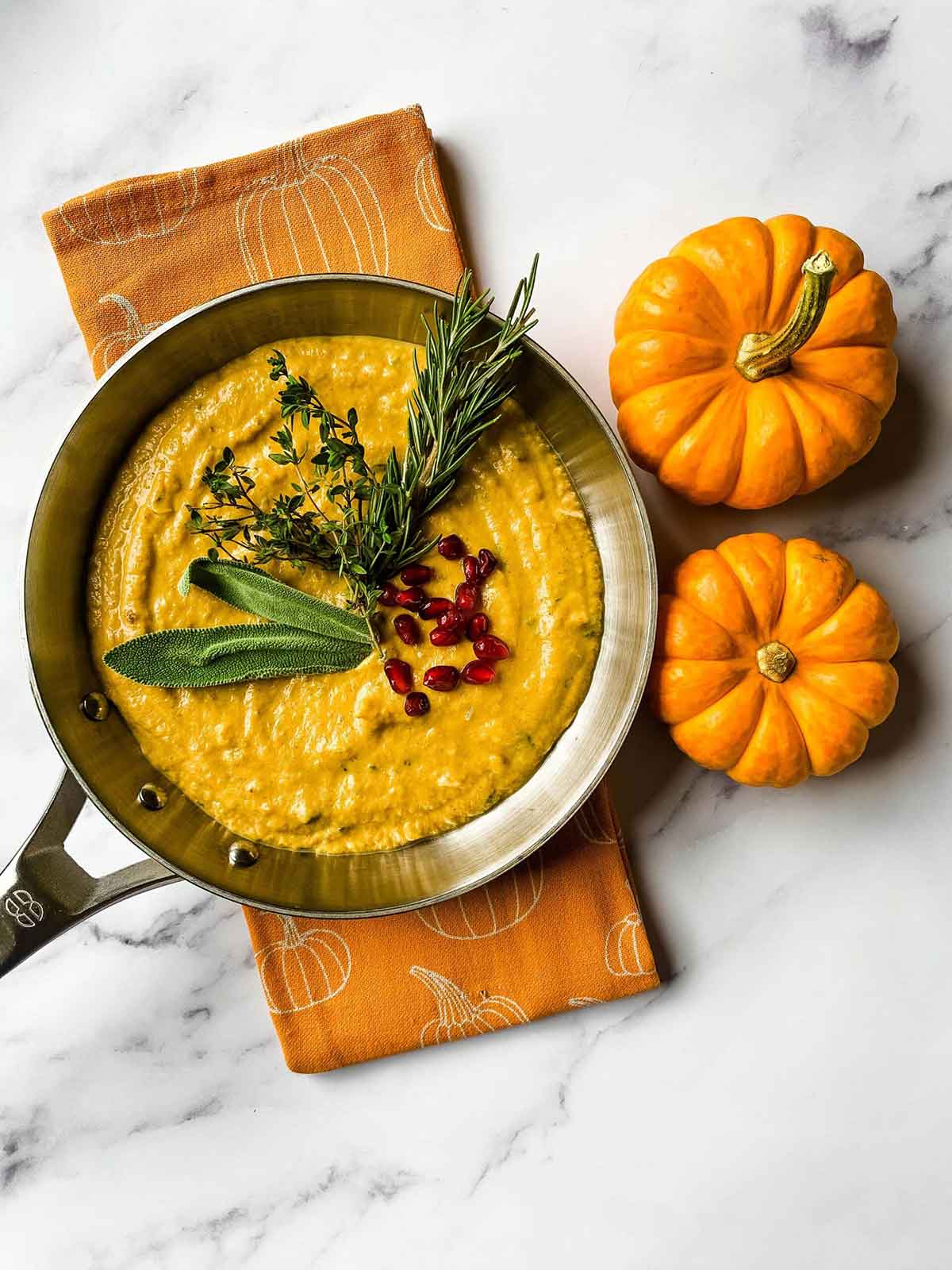 A pan of pumpkin cream sauce on a table beside 2 small pumpkins.