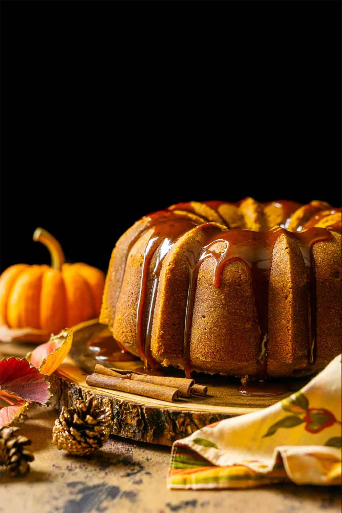 A pumpkin cake on a wooden table beside a small pumpkin and scattered pinecones.