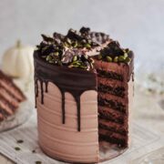 A layered cake, drizzled with chocolate, on a table with a white pumpkin in the background.