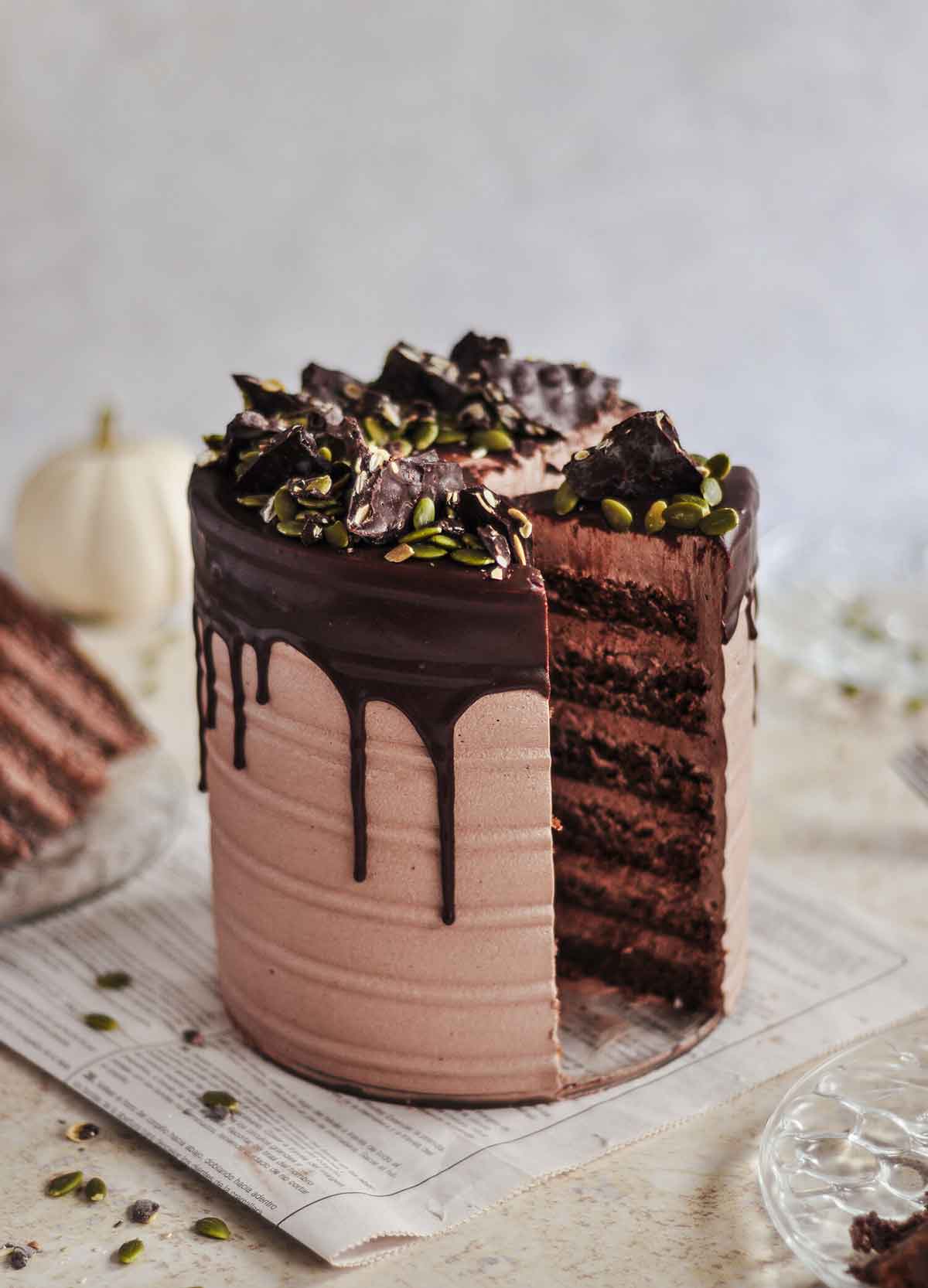 A layered cake, drizzled with chocolate, on a table with a white pumpkin in the background.