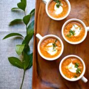 4 cups of pumpkin soup on a wooden tray on a table, beside a sprig of leaves.