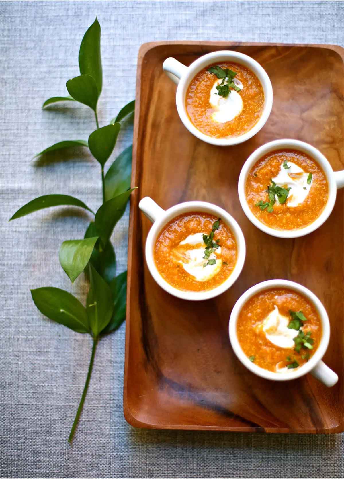 4 cups of pumpkin soup on a wooden tray on a table, beside a sprig of leaves.